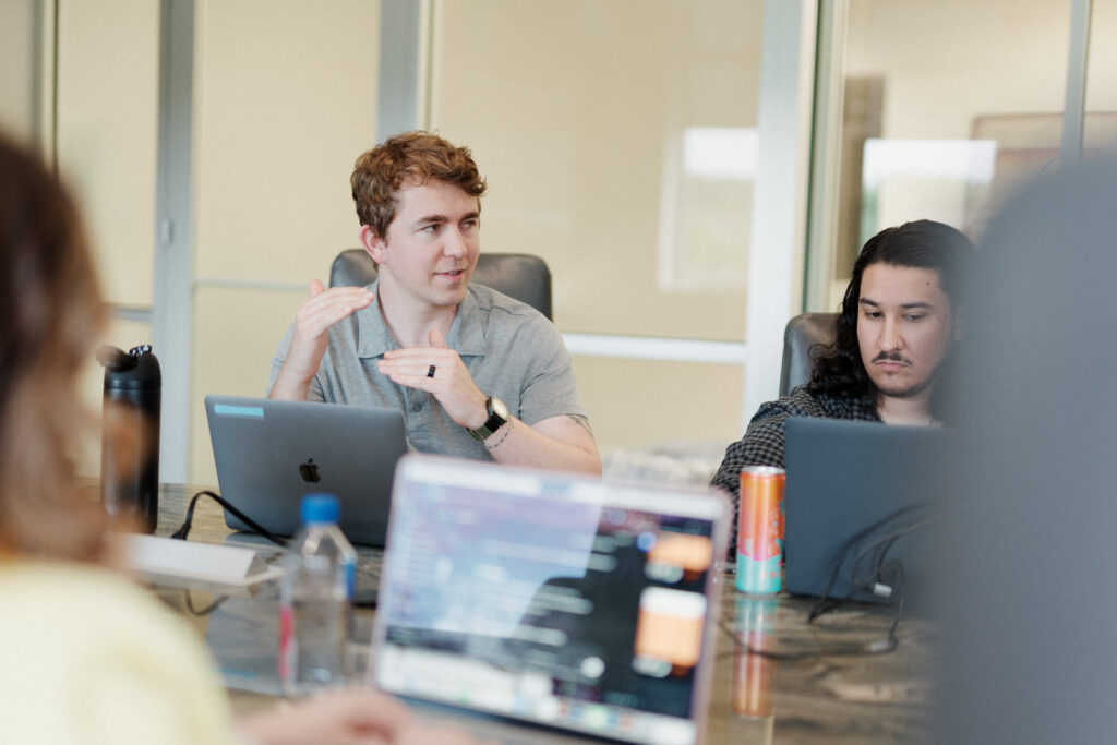 Two employees of a digital ads agency sit at a conference table during a team meeting about results from the ads. One gestures with his hands while speaking, suggesting he’s explaining a concept, while the other focuses intently on his laptop. Open laptops, drinks, and blurred foreground screens indicate an active, collaborative work environment.