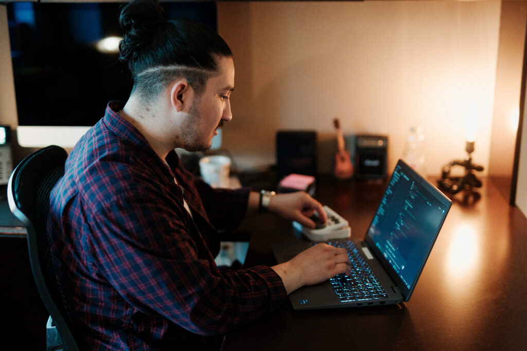 A man with a tied-back hairstyle and plaid shirt works on a laptop at a dimly lit desk, writing code in a development environment. He interacts with a controller beside the keyboard, and the workspace includes a desktop monitor, coffee mug, and decorative items, creating a cozy, tech-focused setup.