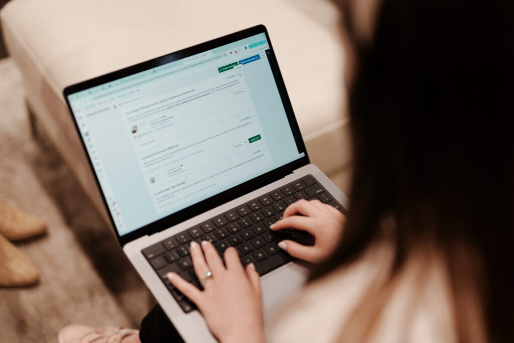A woman types on a laptop while seated in a casual office or lounge setting. The screen displays a web-based content management or collaboration platform, with approval buttons and comment threads visible. Her hands are on the keyboard, suggesting focused work on content creation services.