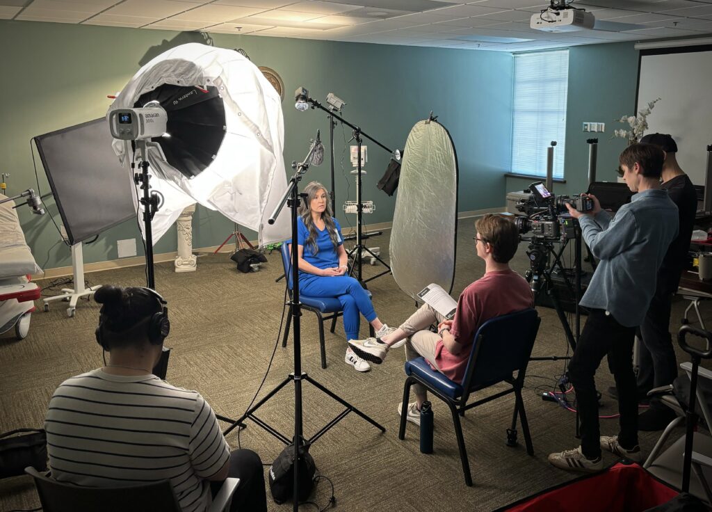A behind-the-scenes look at a professional photo and video agency shoot in a medical office setting. A woman in blue scrubs sits in front of studio lights, cameras, and microphones while a production team records her. Crew members operate cameras, monitor audio, and adjust lighting equipment, creating a well-coordinated filming environment.